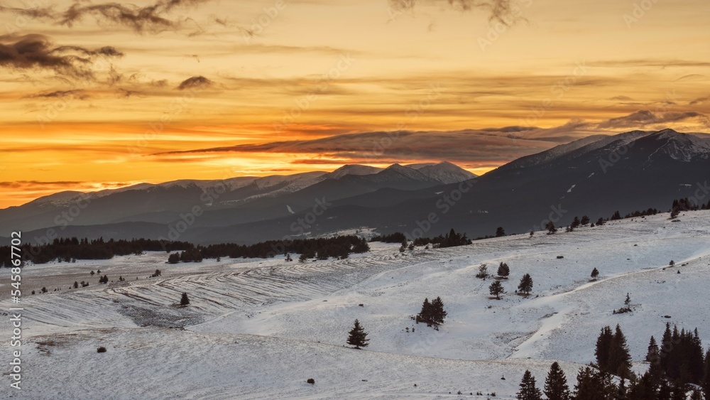 Sunset over snowy peaks in the mountains. Attractive orange cloudy sky, white slopes, natural landscape panoramic view. Perfect winter conditions for travel, sport, recreation and tourism.