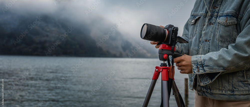 Close-up shot of a photo camera on a tripod and the hands of a woman photographer making settings for shooting on the shore of a foggy lake