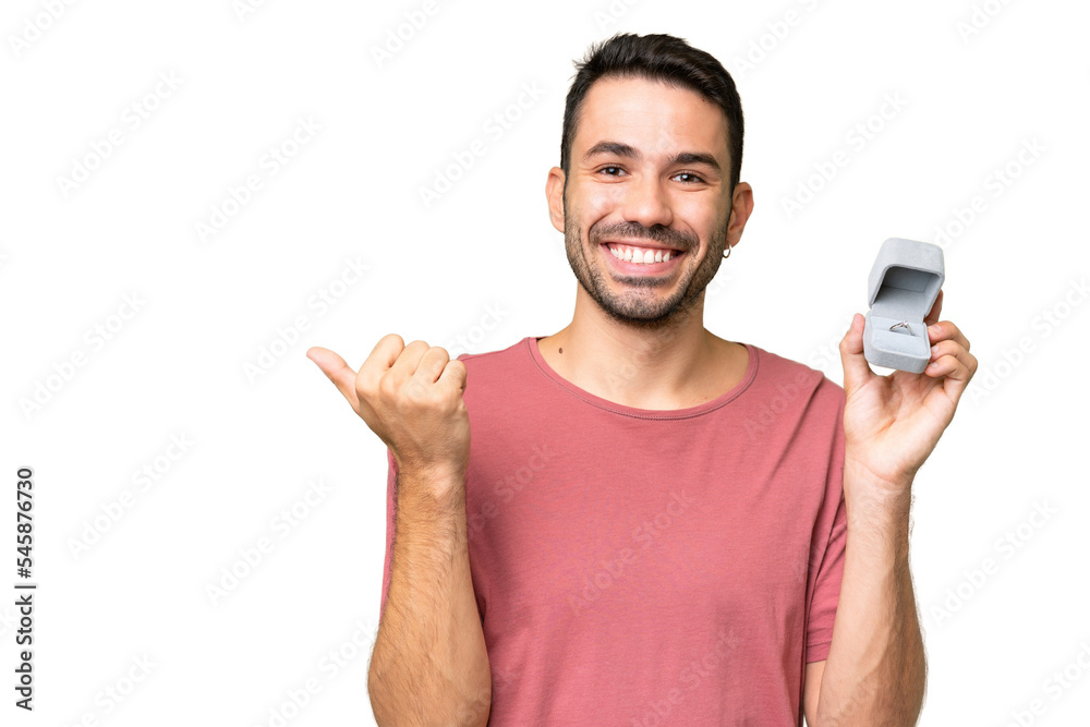 Young handsome caucasian man holding a engagement ring over isolated background pointing to the side to present a product Young handsome caucasian man holding a engagement ring over isolated background pointing to the side to present a product