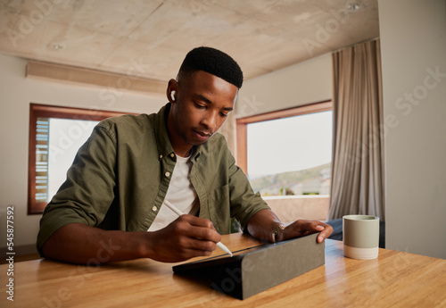 Focussed young adult black African American male writing with pen on electronic tablet while working remotely from apartment. Illustrating and drawing digitally.