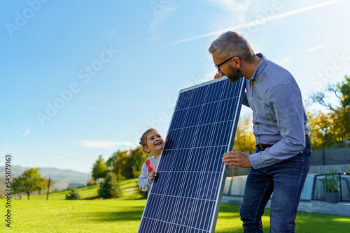 Father with his little daughter catching sun at solar panel,charging at their backyard. Alternative energy, saving resources and sustainable lifestyle concept.