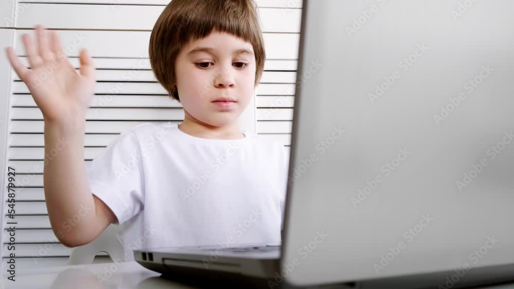 The 5 year child greets by waving his hand while looking at the laptop screen. Online learning and communication over the Internet. Internet scammers and criminals with kid. Home distance e-learning. The 5 year child greets by waving his hand while looking at the laptop screen. Online learning and communication over the Internet. Internet scammers and criminals with kid. Home distance e-learning.
