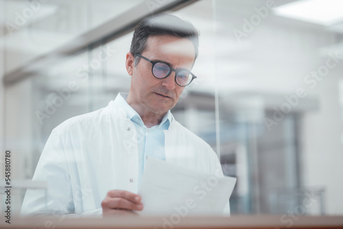 Man in lab coat analyzing medical reports in laboratory seen through glass photo