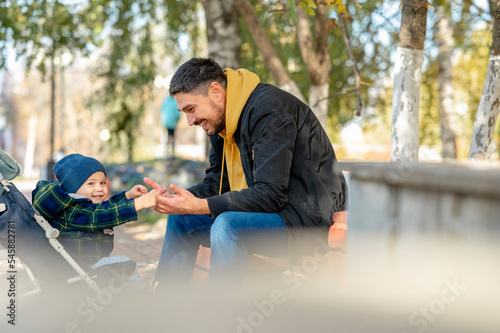 Happy baby boy in stroller holding hands of father sitting under tree at park photo