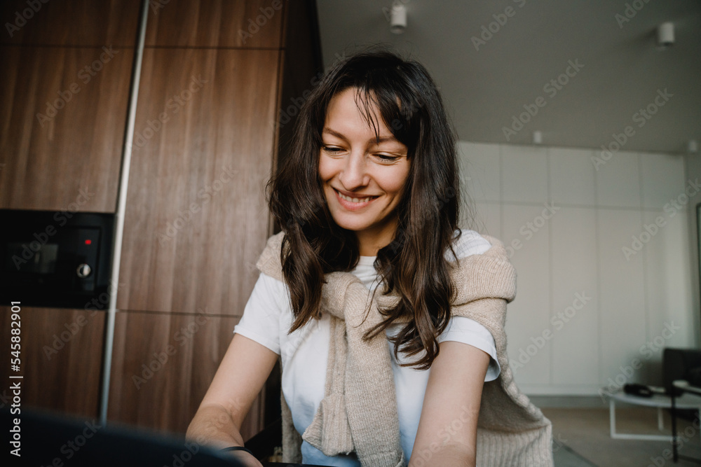 Young white happy woman smiling and sitting with laptop by table at home Young white happy woman smiling and sitting with laptop by table at home