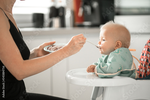 Mother spoon feeding her baby boy child in baby chair with fruit puree in kitchen at home. Baby solid food introduction concept.