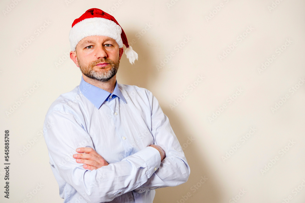Man in light blue shirt and red Christmas hat. Hands crossed. Light background. Celebrating New Year concept. Male at a party. The model in his 40s with grey beard and slim athletic body. Man in light blue shirt and red Christmas hat. Hands crossed. Light background. Celebrating New Year concept. Male at a party. The model in his 40s with grey beard and slim athletic body.