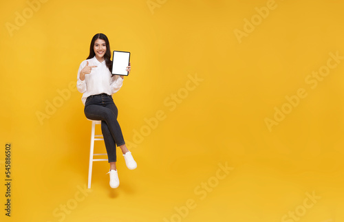 Happy young asian woman showing empty tablet screen while sitting on white chair on yellow studio background. full body length. Excited lady recommending nice and useful tablet application
