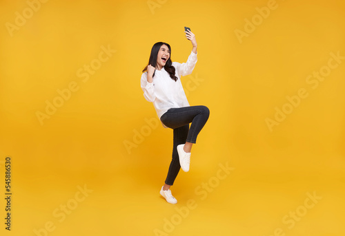 Obraz na plátně Happy young asian teen woman celebrating with mobile phone isolated over yellow background