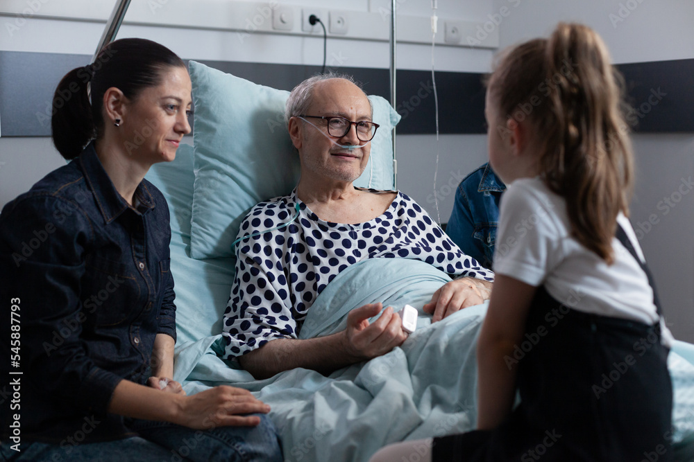 Elderly man lying in hospital bed chatting with visiting family members. Little girl talking to grandfather in elderly people medical clinic. Family living with old man resting in sanatorium bed. - obrazy, fototapety, plakaty 