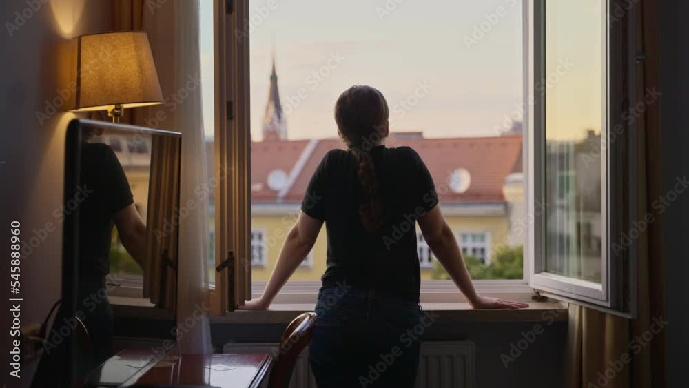 Young woman standing at hotel room or apartment building window looking outside. Female on vacation or holiday enjoying the view of European country in the afternoon. Relaxing looking around the city Young woman standing at hotel room or apartment building window looking outside. Female on vacation or holiday enjoying the view of European country in the afternoon. Relaxing looking around the city