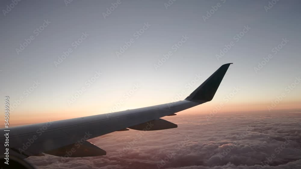 View out window of commercial airline plane wing with soft orange and magenta hues at sunset with clouds below View out window of commercial airline plane wing with soft orange and magenta hues at sunset with clouds below