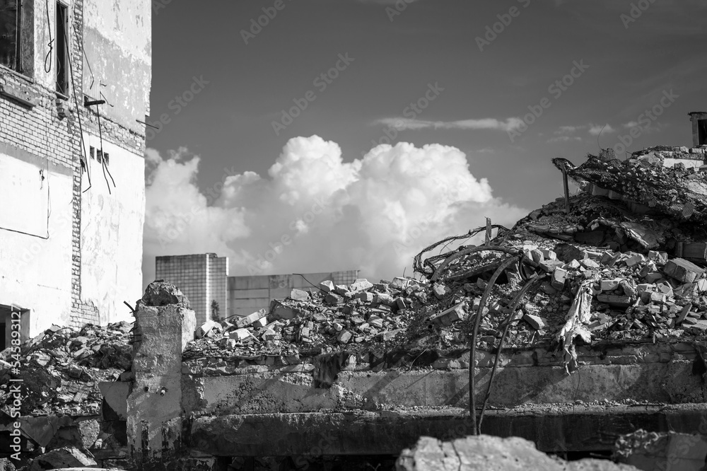 Destruction in the form of a pile of concrete debris against the background of the remains of a house and clouds. Black and white background