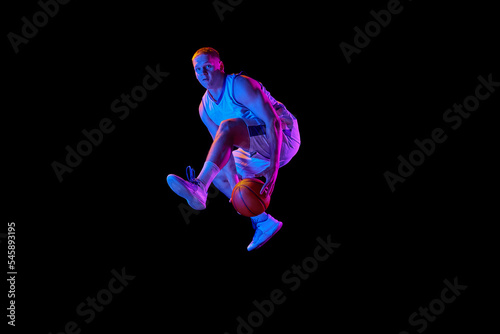 Dynamic portrait of young active athlete, male basketball player in sports uniform practicing with ball, jumping, dribbling isolated over dark background in neon light