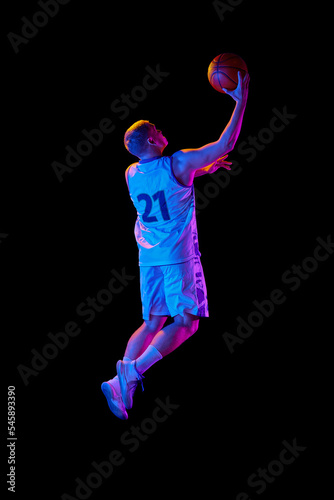 Studio shot of young active athlete, male basketball player in sports uniform in motion and action with ball isolated over dark background in neon light
