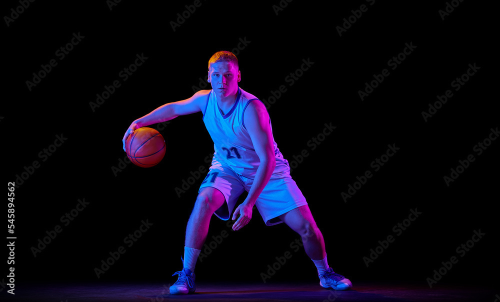 Fototapeta premium Dynamic portrait of young active athlete, male basketball player in sports uniform practicing with ball, jumping, dribbling isolated over dark background in neon light