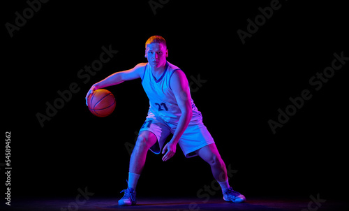 Dynamic portrait of young active athlete, male basketball player in sports uniform practicing with ball, jumping, dribbling isolated over dark background in neon light © master1305