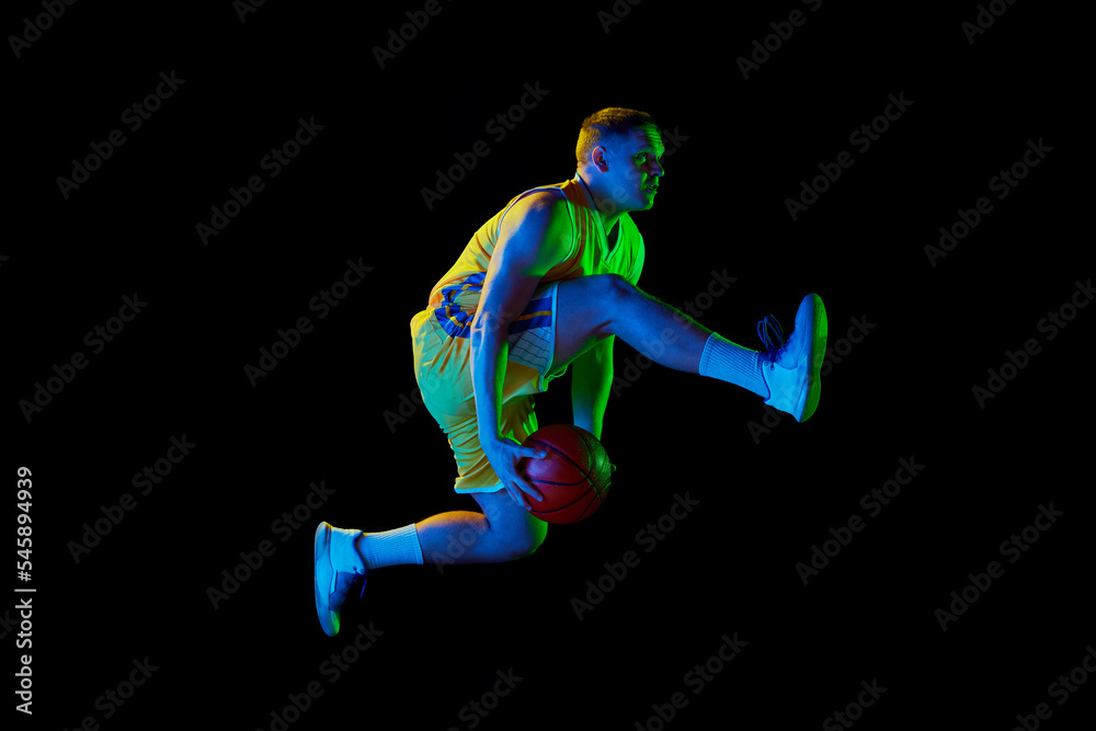 Studio shot of young active athlete, male basketball player in sports uniform in motion and action with ball isolated over dark background in neon light