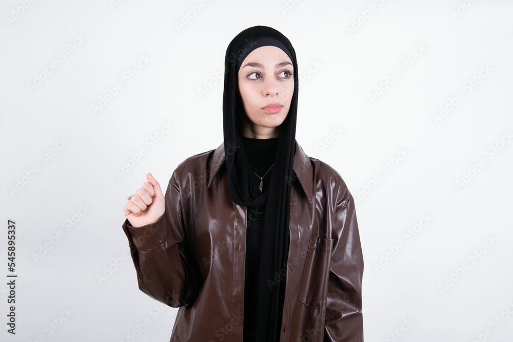 young beautiful muslim woman wearing hijab and leather jacket over white background, looks pensively aside, plans actions after university, imagines what to do Thinks over about new project.