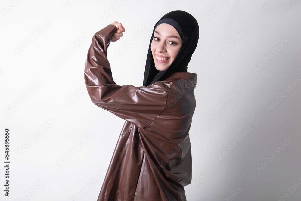 young beautiful muslim woman wearing hijab and leather jacket over white background,  showing muscles after workout. Health and strength concept.