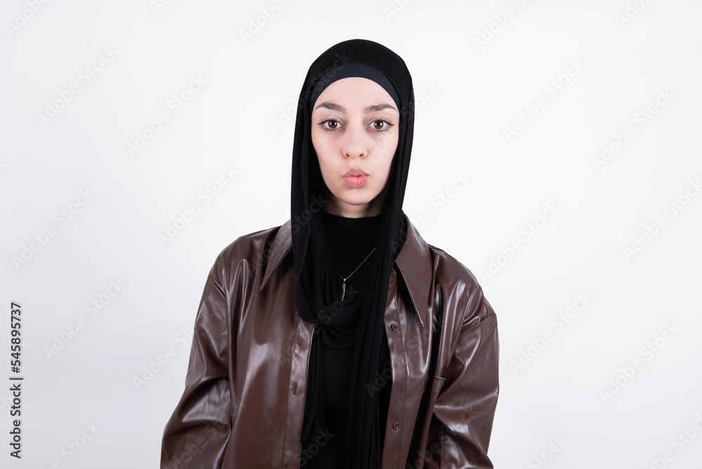 young beautiful muslim woman wearing hijab and leather jacket over white background crosses eyes, puts lips, makes grimace with awkward expression has fun alone, plays fool.