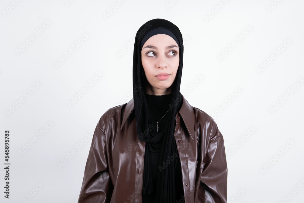 young beautiful muslim woman wearing hijab and leather jacket over white background has worried face looking up lips together, being upset thinking about something important, keeps hands down.