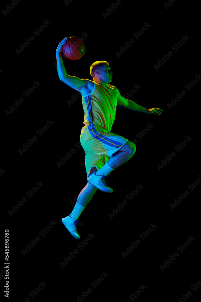 Dynamic portrait of young active athlete, male basketball player in sports uniform practicing with ball, jumping, dribbling isolated over dark background in neon light