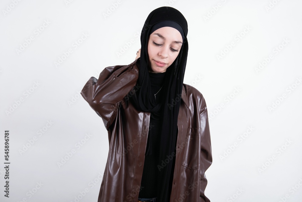 young beautiful muslim woman wearing hijab and leather jacket over white background suffering from back and neck ache injury, touching neck with hand, muscular pain.