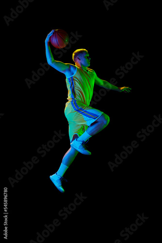 Dynamic portrait of young active athlete  male basketball player in sports uniform practicing with ball  jumping  dribbling isolated over dark background in neon light