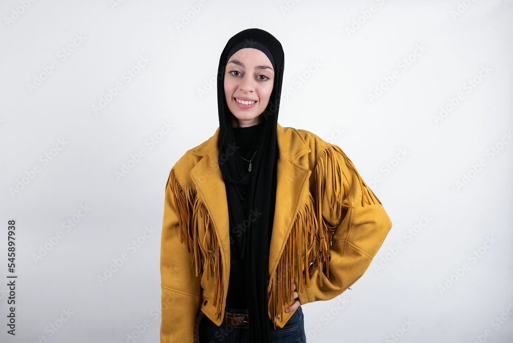 Studio shot of cheerful young beautiful muslim woman wearing hijab and yellow jacket over white background keeps hand on hip, smiles broadly. Studio shot of cheerful young beautiful muslim woman wearing hijab and yellow jacket over white background keeps hand on hip, smiles broadly.
