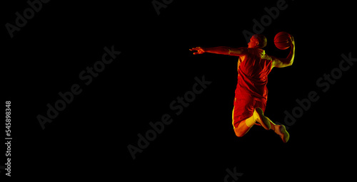 Young athletic male basketball player jumping with basketball ball isolated over dark background in red neon light. Concept of energy, professional sport, hobby.