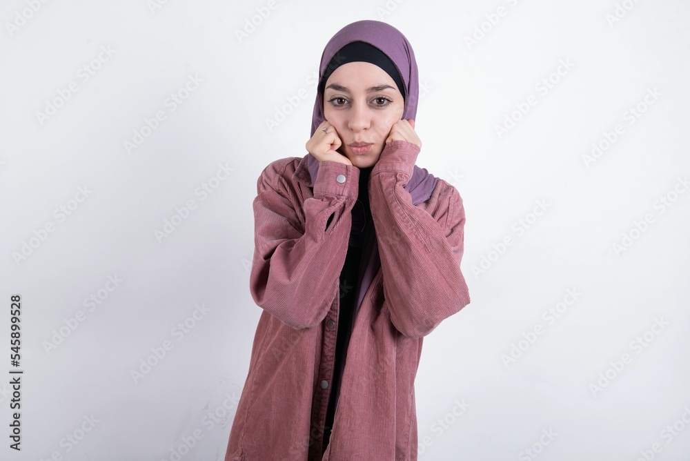young beautiful muslim woman wearing hijab and pink jacket over white background with surprised expression keeps hands under chin keeps lips folded makes funny grimace