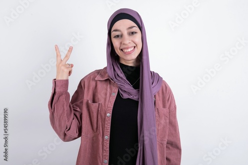 young beautiful muslim woman wearing hijab and pink jacket over white background smiling and looking friendly, showing number two or second with hand forward, counting down © Jihan