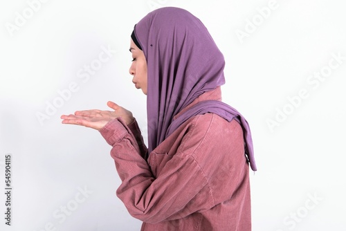 Profile side view view portrait of attractive young beautiful muslim woman wearing hijab and pink jacket over white background sending air kiss