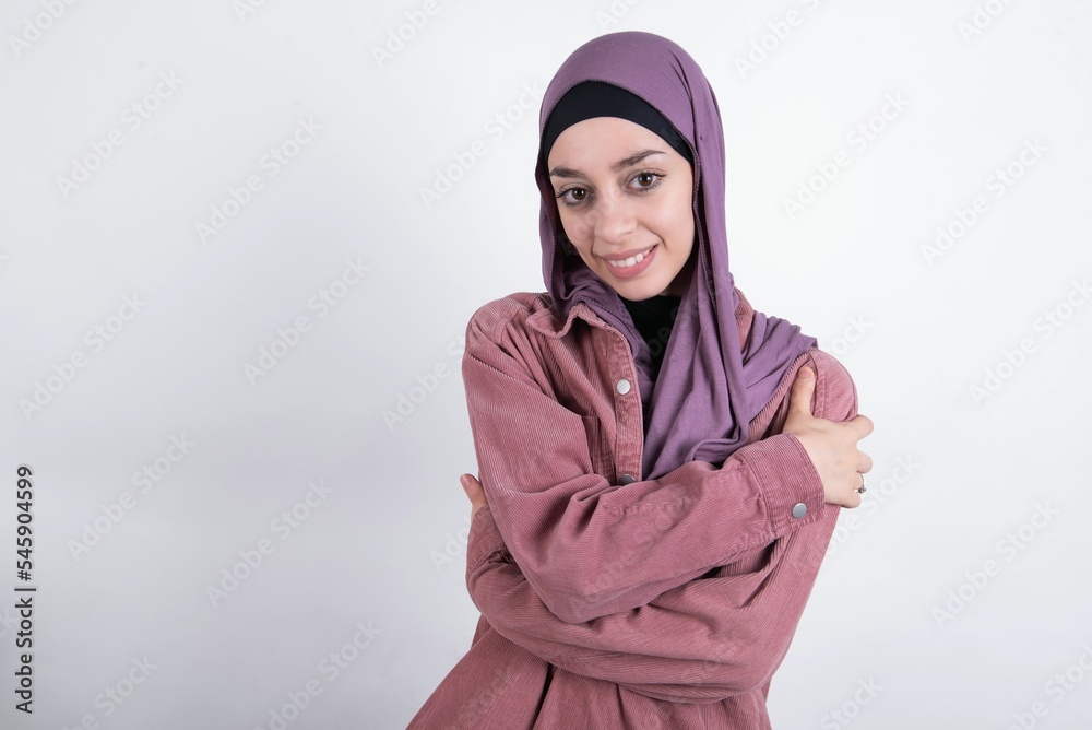 Charming pleased young beautiful muslim woman wearing hijab against white background embraces own body, pleasantly feels comfortable poses. Tenderness and self esteem concept Charming pleased young beautiful muslim woman wearing hijab against white background embraces own body, pleasantly feels comfortable poses. Tenderness and self esteem concept