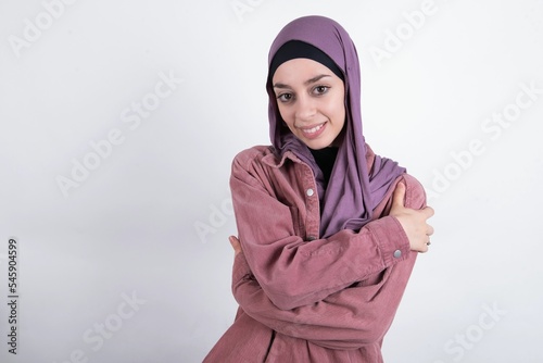 Charming pleased young beautiful muslim woman wearing hijab against white background embraces own body, pleasantly feels comfortable poses. Tenderness and self esteem concept photo