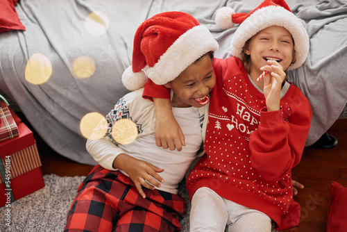 Happy interracial boys wearing festive costumes, Santa Claus hats, licking candy canes, having fun