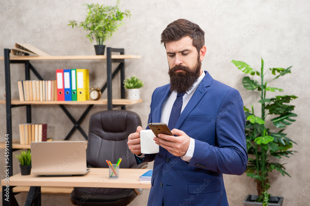 Serious manager in suit drinking coffee while reading text message on smartphone in office, SMS Serious manager in suit drinking coffee while reading text message on smartphone in office, SMS