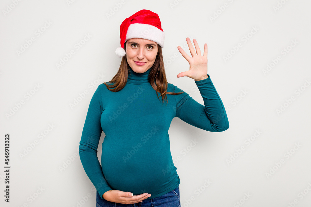 Pregnant caucasian woman wearing a christmas hat isolated on white background smiling cheerful showing number five with fingers. Pregnant caucasian woman wearing a christmas hat isolated on white background smiling cheerful showing number five with fingers.