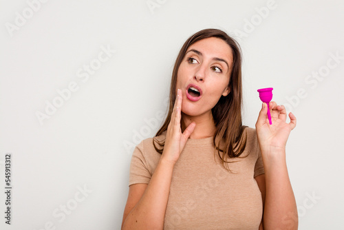 Young caucasian woman holding menstrual cup isolated on white background is saying a secret hot braking news and looking aside