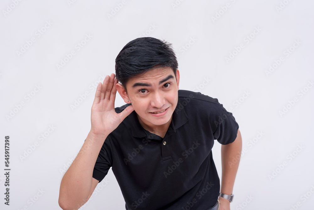 A young asian man leans forward to hear a juicy rumor. Having difficulty hearing, partially deaf, or trying to listen to inaudible speech. Isolated on a white background.