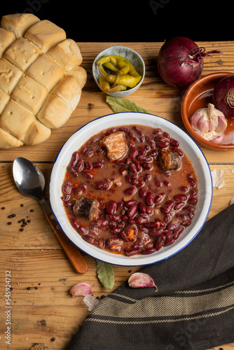 Aerial view of a plate of Basque beans on a rustic wooden table with a spoon, onions, garlic and a loaf of bread, vertical photo