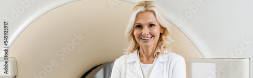 middle aged radiologist smiling at camera near computed tomography machine, banner.