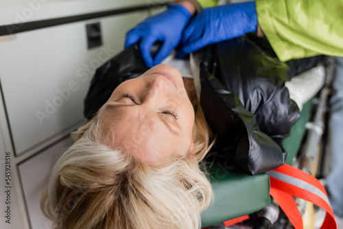 Blurred paramedic taking off jacket from unconscious middle aged woman during first air in emergency vehicle.