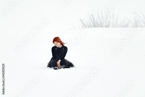 A sad redhead girl sits in the snow in winter in a large white empty space