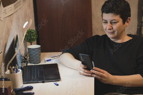 Fotografie Middle-aged brunette woman sits at a desk in front of laptop screen and looks at smartphone screen