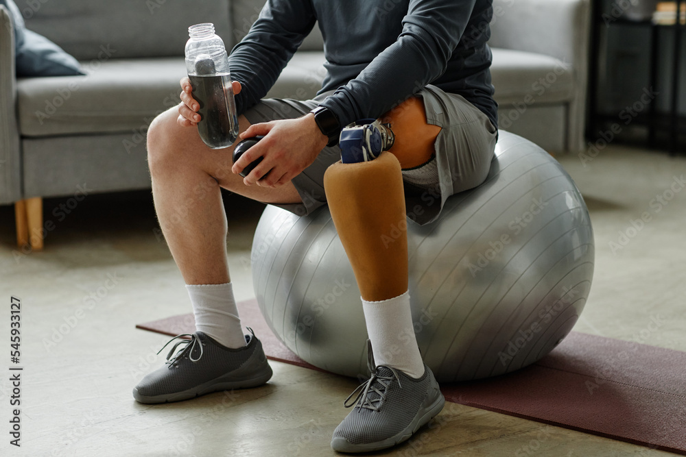 Close up of unrecognizable man with prosthetic leg sitting on fitness ball and holding water bottle after home workout