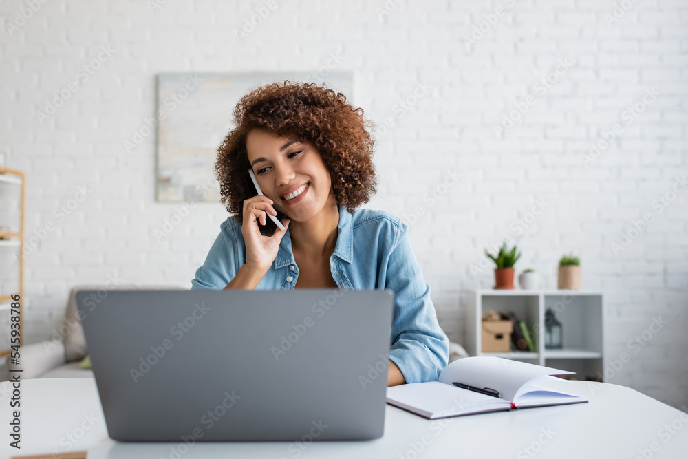 Positive african american freelancer talking on cellphone and looking at laptop in living room. Positive african american freelancer talking on cellphone and looking at laptop in living room.