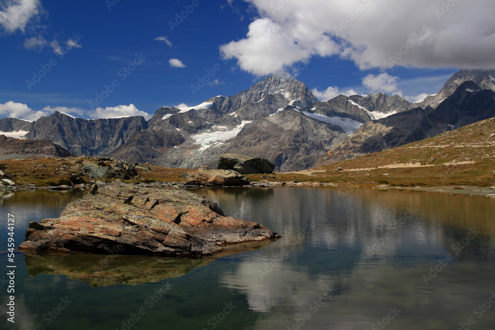 Landscape with a smooth surface of the lake Riffelsee, mountains and clouds reflected in it, with a large stone on the foreground, on a mountain Gornergrat, near Zermatt, in southern Switzerland