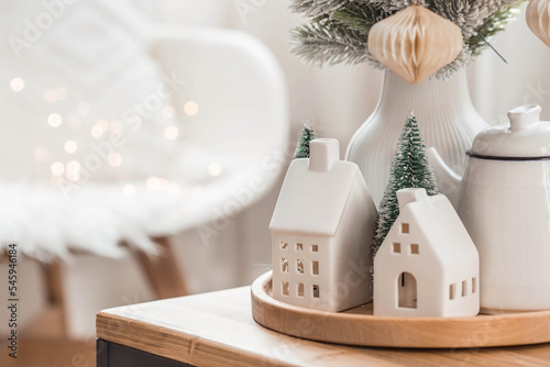 A bouquet of fir trees, a plaid in a wicker basket and Scandinavian white houses on a wooden table in the home interior of the living room. A cozy concept of festive home decoration.